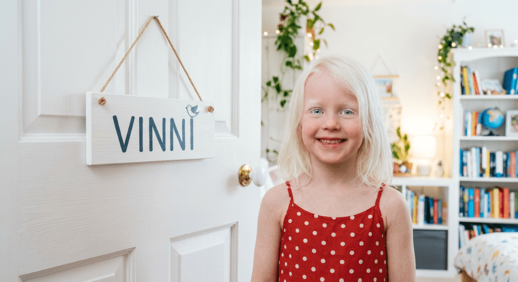 Smiling child standing indoors near a wooden door with a sign that reads VINNI